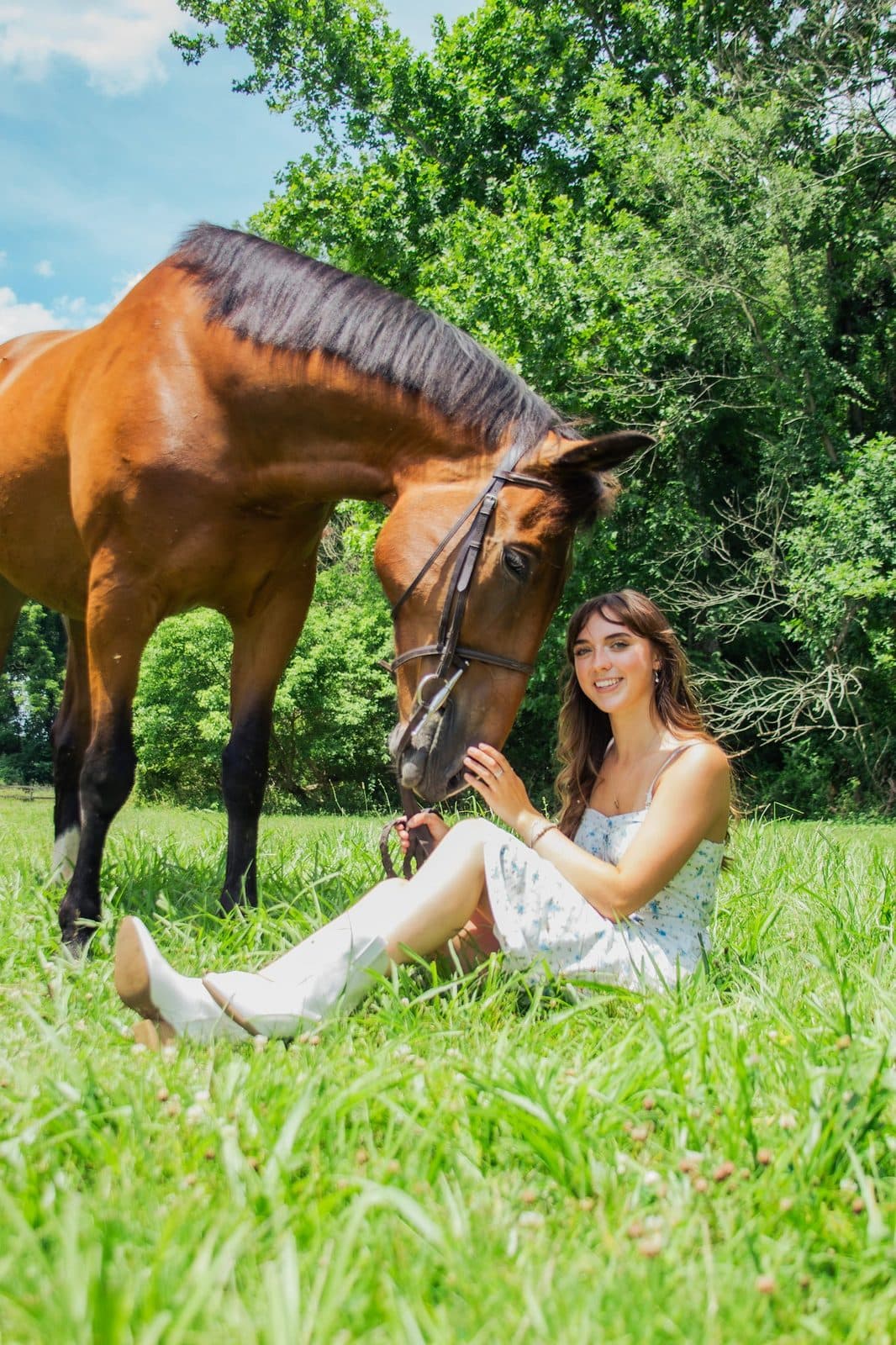 A bay horse lowering its head to Aubrey Russo as she sits in a grassy field