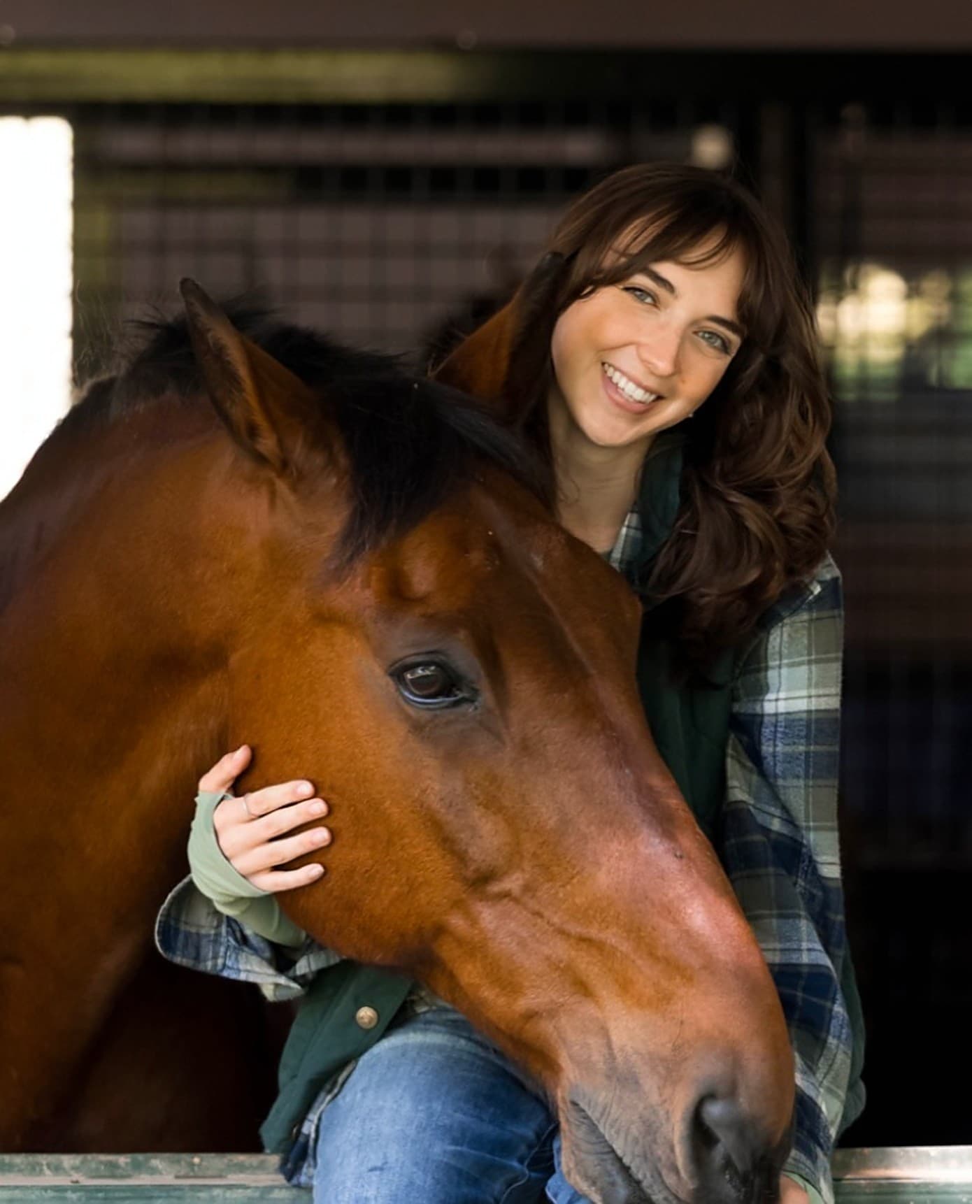 Aubrey Russo smiling beside a bay horse resting its head on her at a stall door