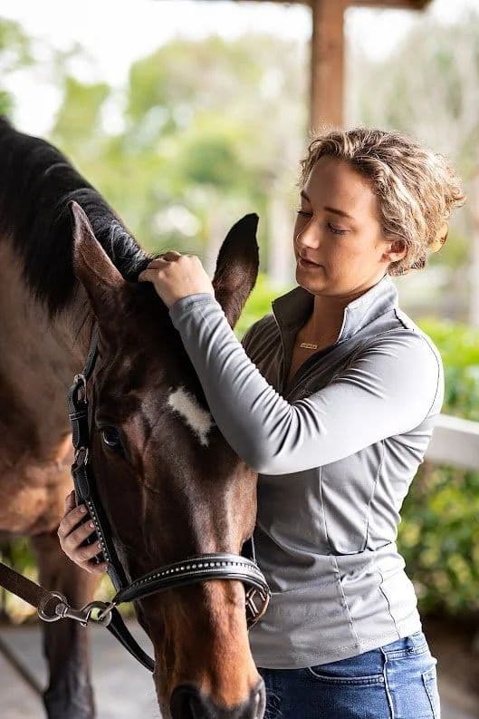 Maija Luttinger standing beside a bay horse in the barn aisle